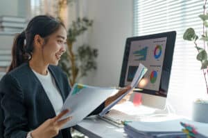Young office worker smiling while analyzing financial charts and graphs, holding documents and comparing information with computer screen, working with statistics and reports in modern office